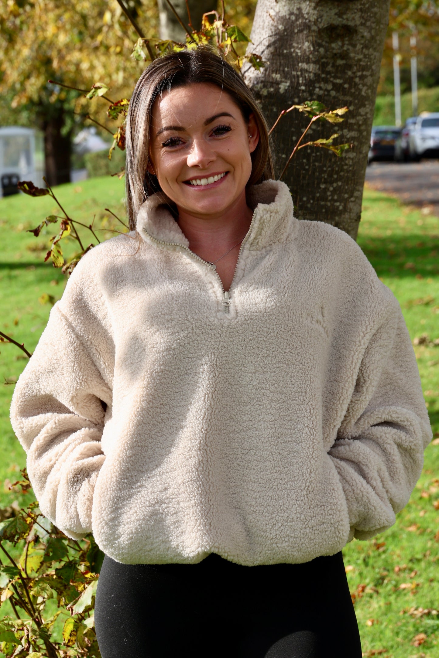 Woman wearing a beige fleece pullover standing outdoors with trees and grass in the background